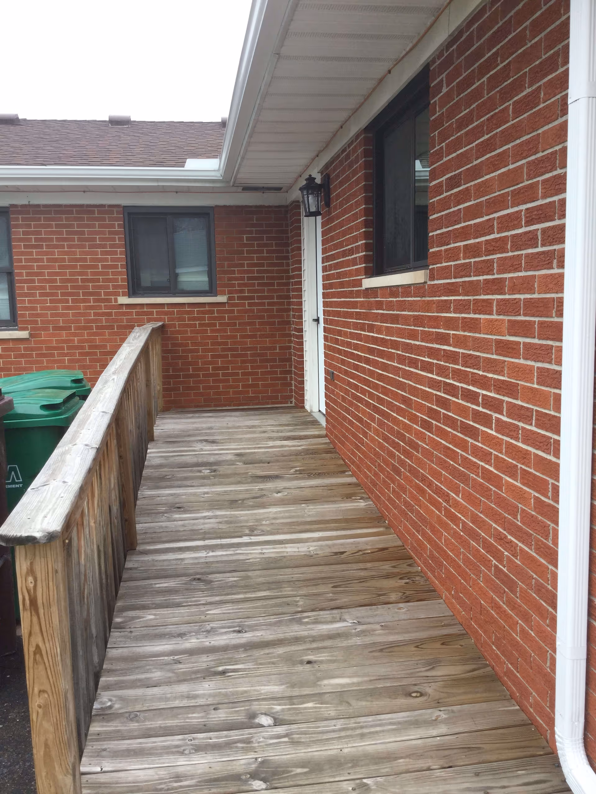 A wooden ramp with a handrail leading to a white door on a red brick building. There are two windows visible on the brick walls and a black outdoor light fixture mounted near the door. Two green trash bins are partially visible on the left side near the ramp.