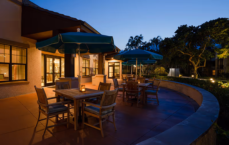 Outdoor patio area at dusk with wooden tables and chairs under green umbrellas, adjacent to a building with lit windows and surrounded by trees and landscaping.