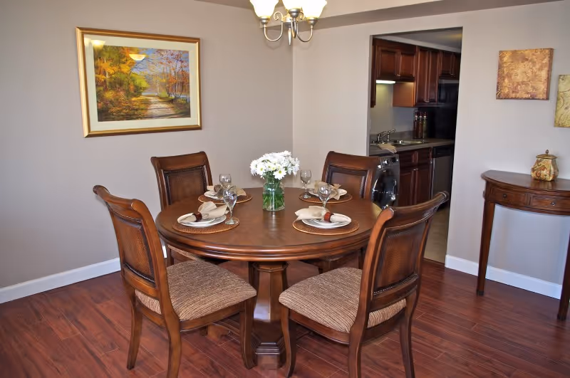 A dining room with a round wooden table set for four with plates, napkins, and wine glasses. A vase with white flowers is in the center of the table. Four wooden chairs with cushioned seats surround the table. The room has wooden flooring, beige walls, a chandelier overhead, and framed artwork on the walls. A small wooden side table is against the wall, and a kitchen area is visible through an open doorway.
