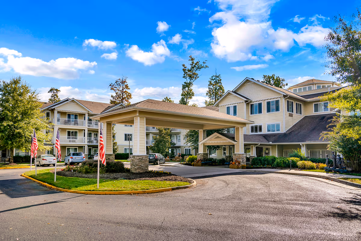 Exterior view of Chesterfield Heights Gracious Retirement Living facility showing a large covered entrance with stone pillars, multiple American flags, parked cars, and a multi-story building surrounded by trees under a partly cloudy blue sky.