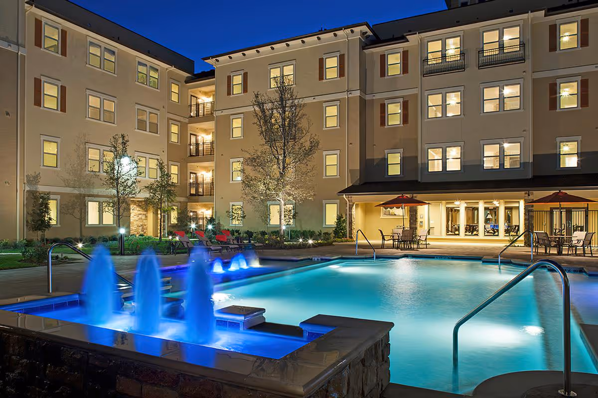 Night view of an illuminated outdoor swimming pool with blue water fountains in front of a multi-story senior living facility building with many lit windows and outdoor seating areas with tables and umbrellas.
