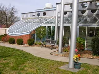 Outdoor view of a facility with a glass sunroom attached to a brick wall, a concrete walkway, a bench, some bushes, and a small flower pot near metal poles.
