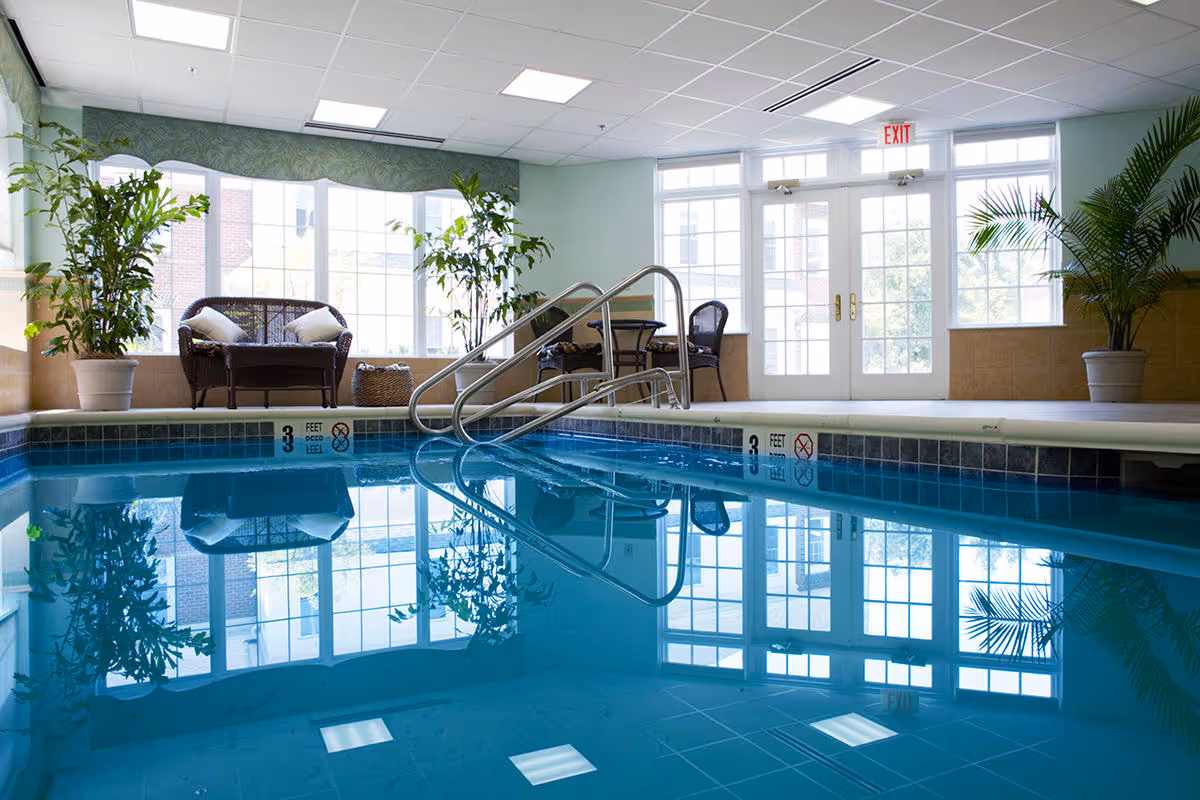 Indoor swimming pool area with clear blue water, metal handrails, and a seating area with wicker chairs and potted plants near large windows and glass doors letting in natural light.