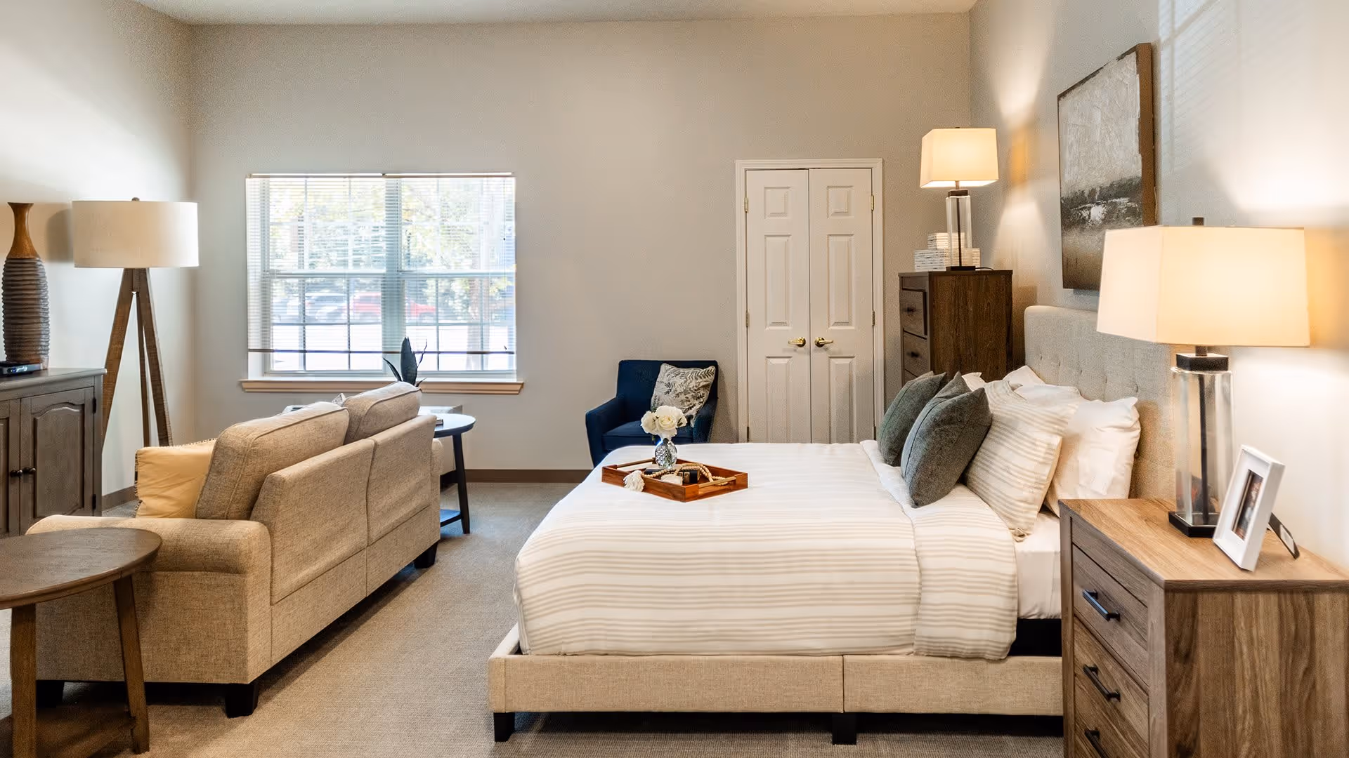 A well-lit bedroom featuring a neatly made bed with striped bedding and multiple pillows. To the right of the bed is a wooden nightstand with a lamp and a framed photo. Behind the bed is a dresser with a lamp and decorative items. A blue armchair is positioned near a window with blinds, and a beige sofa with cushions is placed opposite the bed. The room has neutral-colored walls and carpeted flooring.