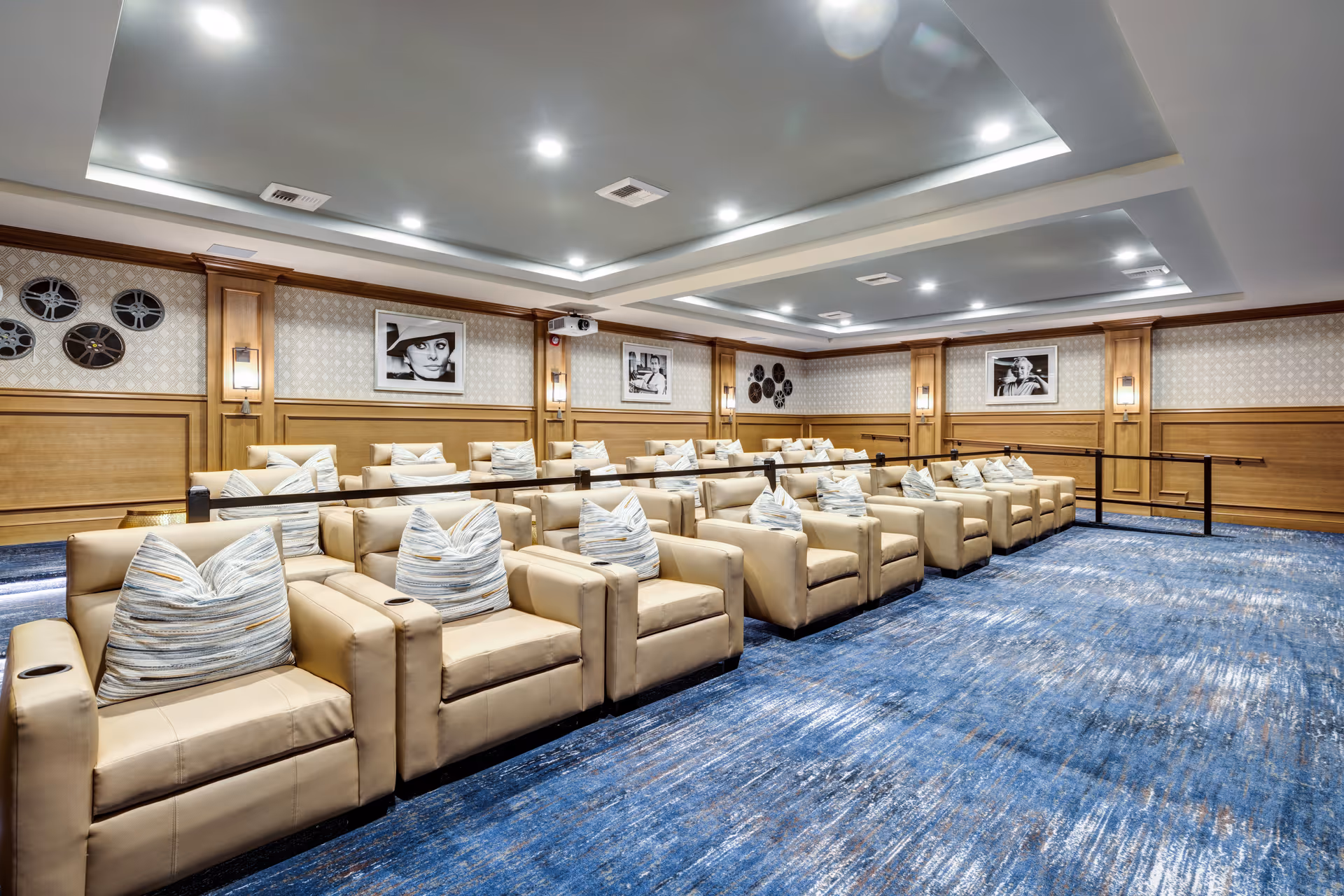 A modern theater room with multiple rows of beige leather recliner chairs, each with a striped pillow. The room features blue patterned carpet, wood-paneled walls with framed black and white photographs, and recessed ceiling lights.