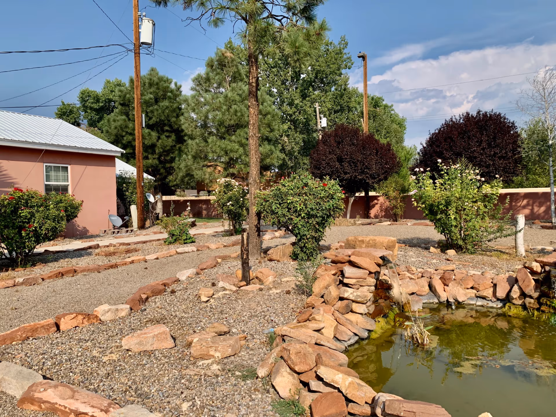 Outdoor garden area with a small pond surrounded by rocks, various bushes, trees, and a gravel pathway. A building with a metal roof and a window is visible on the left side, with utility poles and power lines overhead under a partly cloudy sky.