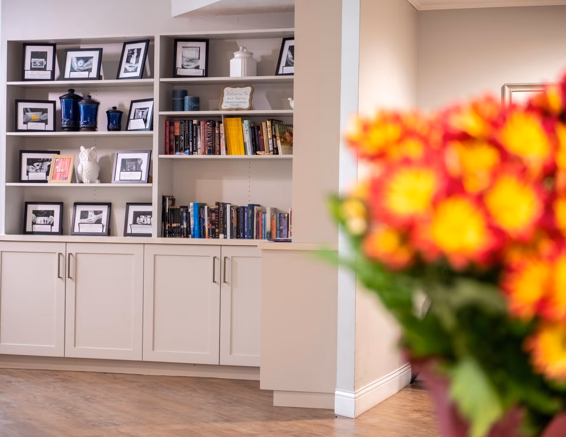 A built-in white bookshelf filled with framed black and white photos, blue decorative jars, a white owl figurine, and various books. In the foreground, there is a blurred bouquet of red and yellow flowers. The room has wooden flooring and light-colored walls.