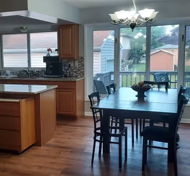 Interior view of a kitchen and dining area with wooden cabinets, a countertop, a coffee maker, and a dining table with six chairs. Large windows and a glass door provide a view of a backyard with a deck, lawn, and neighboring houses.