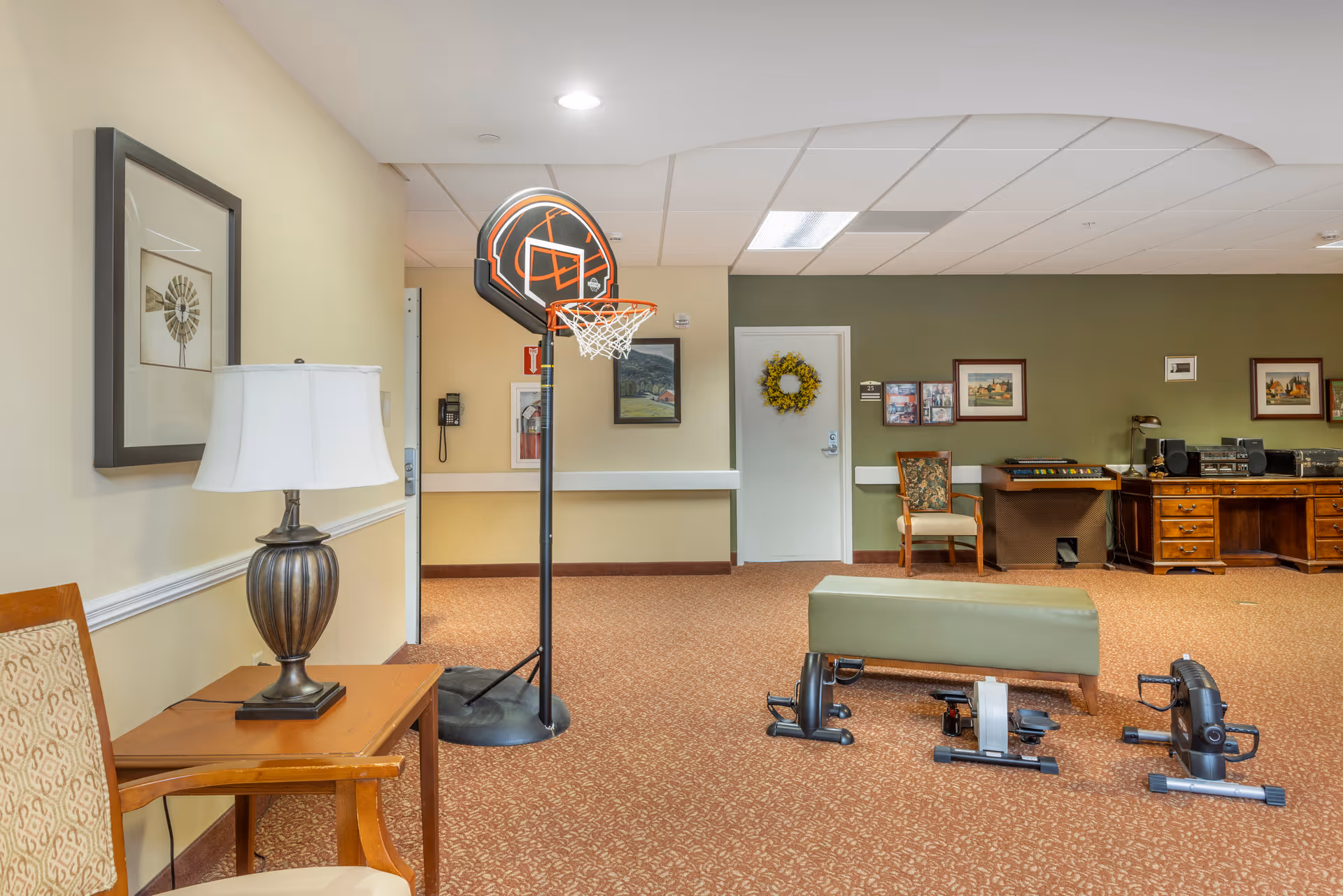 Interior room with beige and green walls, featuring a small basketball hoop, a bench, three pedal exercisers on the carpeted floor, a wooden desk with a stereo system, framed pictures on the walls, and a chair with a side table and lamp.