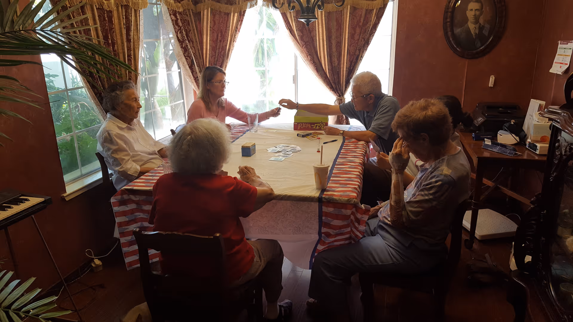 A group of elderly people and a younger woman sitting around a rectangular table covered with a striped tablecloth, playing a card game in a cozy room with large windows and curtains.