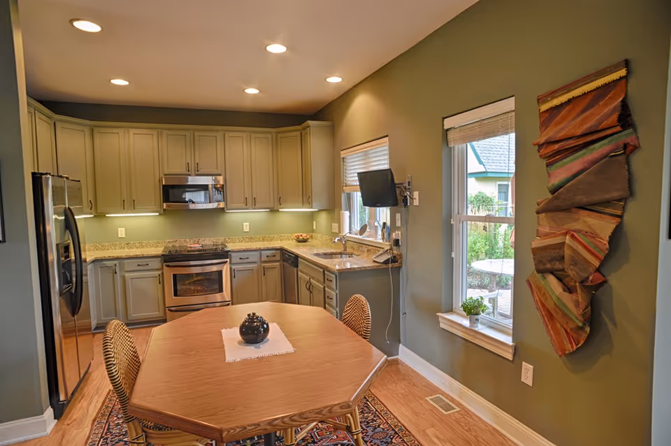 A kitchen with olive green walls, light gray cabinets, granite countertops, and stainless steel appliances including a refrigerator, oven, and microwave. There is a wooden hexagonal table with two wicker chairs in the center on a patterned rug. A small TV is mounted on the wall near two windows with blinds, and a colorful fabric wall hanging is displayed on the right wall.