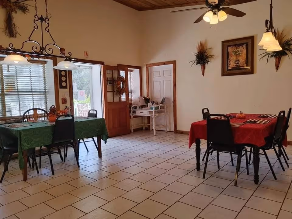 Bright communal dining room with two decorated tables and chairs, tile floor, ceiling fans and wall decorations.