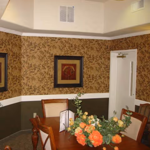 A dining room with a wooden table and chairs. The table has a floral centerpiece with orange and yellow flowers. The walls have a two-tone design with a floral patterned wallpaper on the upper half and a solid dark color on the lower half. There is a framed picture on the wall and a white door in the background.