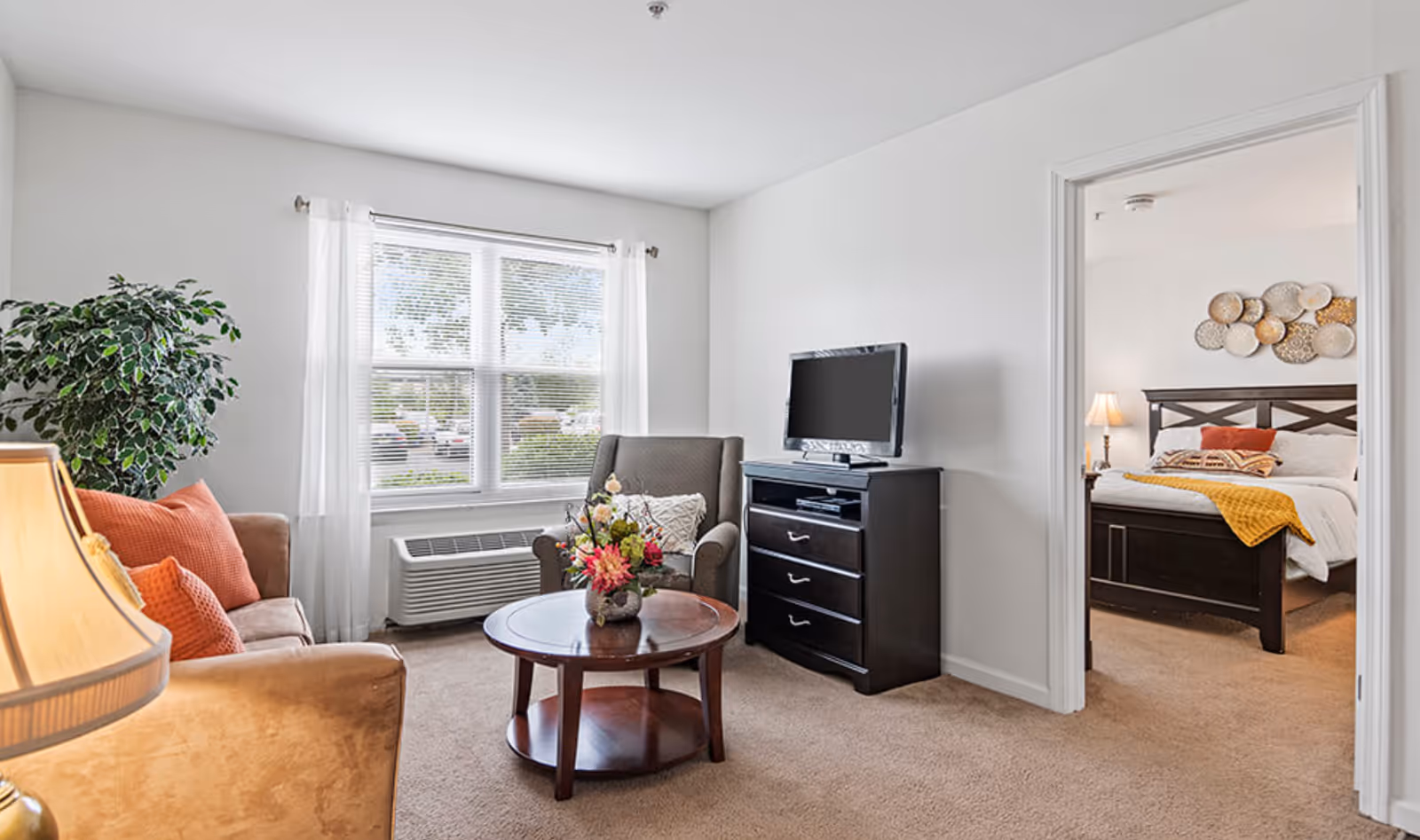 A cozy living room with a beige sofa adorned with orange cushions, a round wooden coffee table with a flower arrangement, a gray armchair, and a TV on a dark wooden dresser. A large window with white curtains lets in natural light. Through an open doorway, a bedroom with a dark wooden bed frame, white bedding, and decorative wall art is visible.