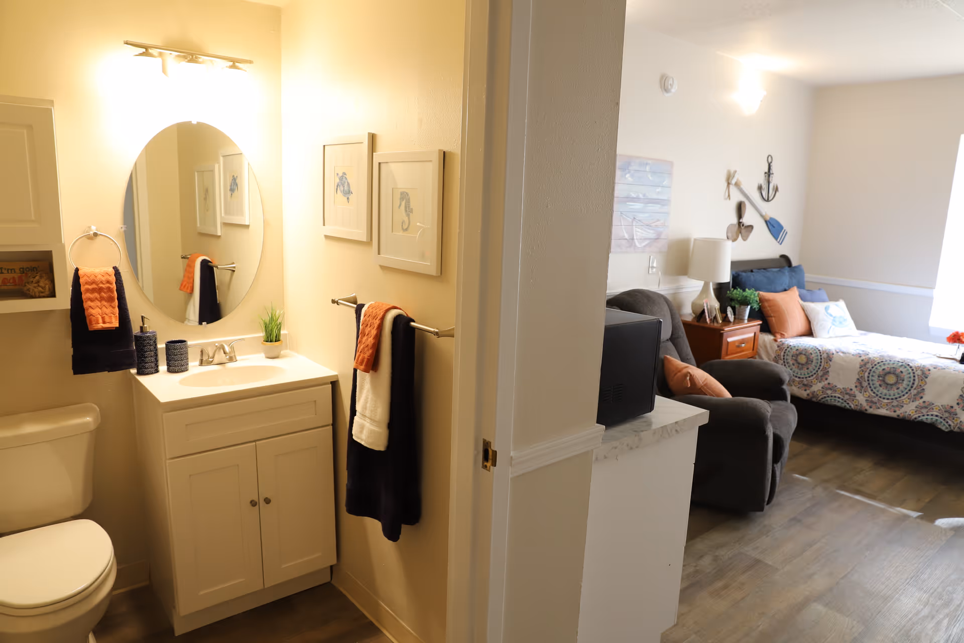 View of a small bathroom and adjacent bedroom in a senior living facility. The bathroom features a white vanity with an oval mirror, a toilet, and towels hanging on racks. The bedroom has a bed with patterned bedding, a recliner chair, a nightstand with a lamp, and nautical-themed wall decorations including an anchor and oars.