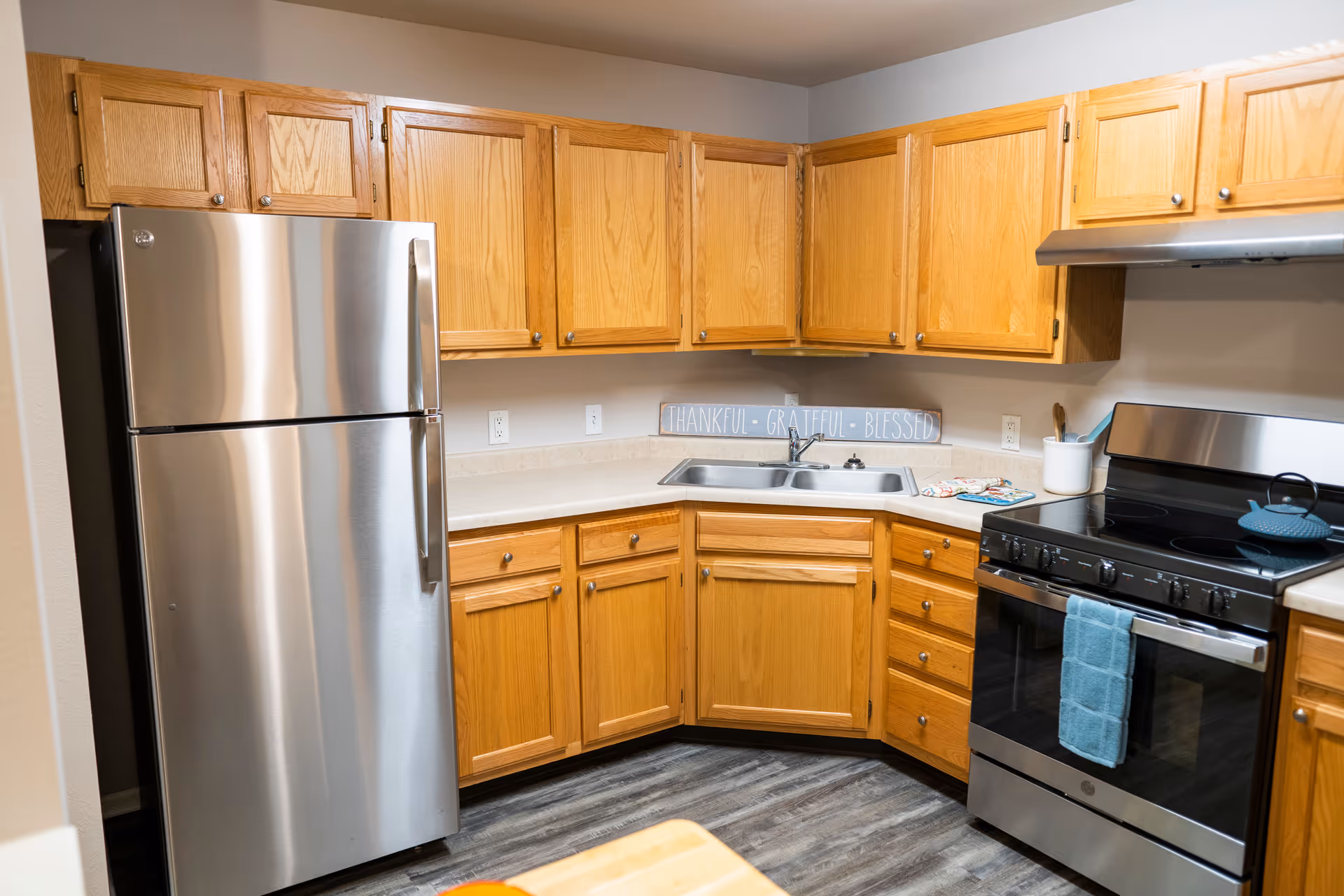 A kitchen with wooden cabinets, a stainless steel refrigerator, a stainless steel stove with a blue towel hanging on the handle, a double sink, and a decorative sign on the countertop that reads 'THANKFUL • GRATEFUL • BLESSED'.