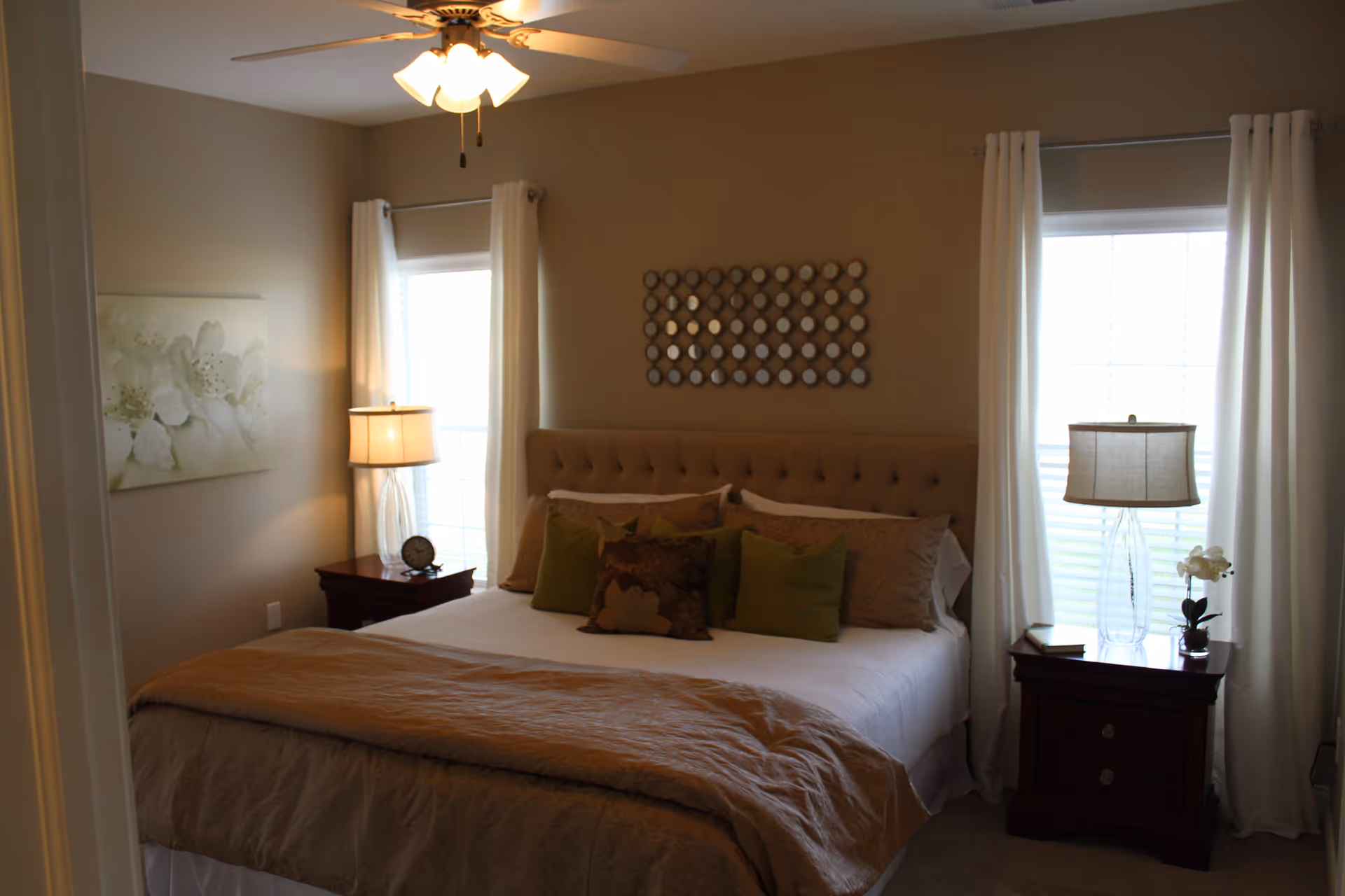 A cozy bedroom with a large bed featuring a tufted headboard, multiple pillows, and a beige blanket. There are two windows with white curtains on either side of the bed, each with a wooden nightstand holding a lamp and decorative items. A ceiling fan with lights is above the bed, and a floral painting and circular mirror wall art decorate the walls.