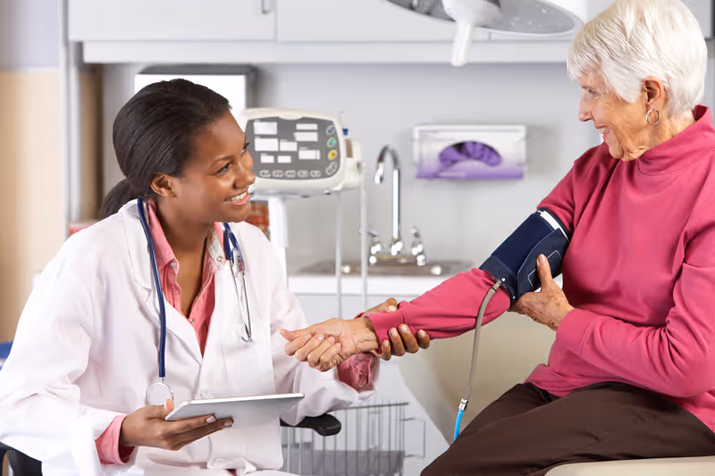A healthcare professional in a white coat is smiling and holding the wrist of an elderly woman wearing a pink sweater while measuring her blood pressure with a cuff in a clinical setting.