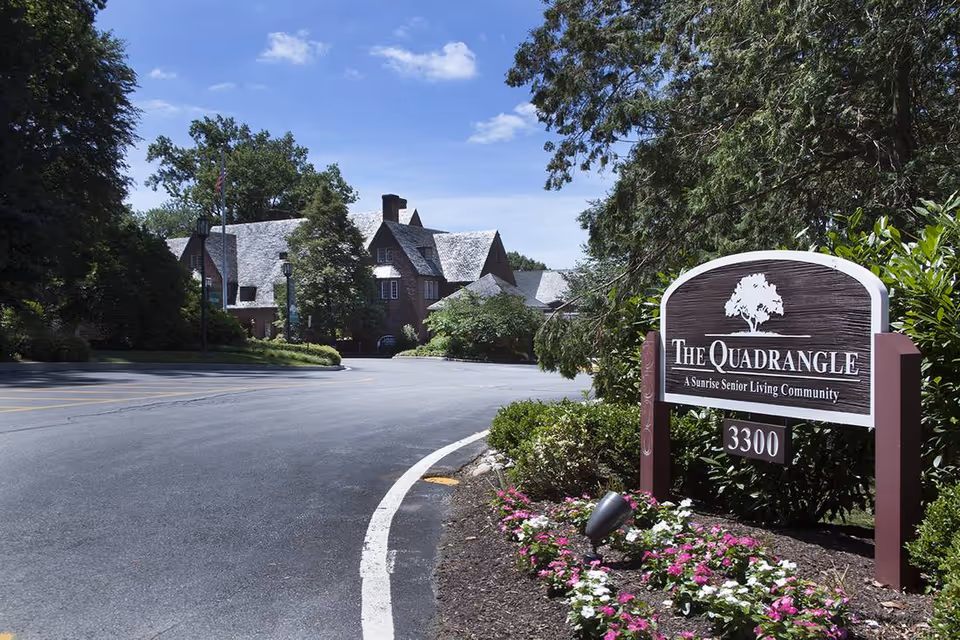 View of the entrance to The Quadrangle senior living community, featuring a curved driveway, landscaped flower beds with pink and white flowers, and a large wooden sign with the community name and address 3300. In the background, there is a large brick building with a steep roof surrounded by trees under a blue sky with some clouds.
