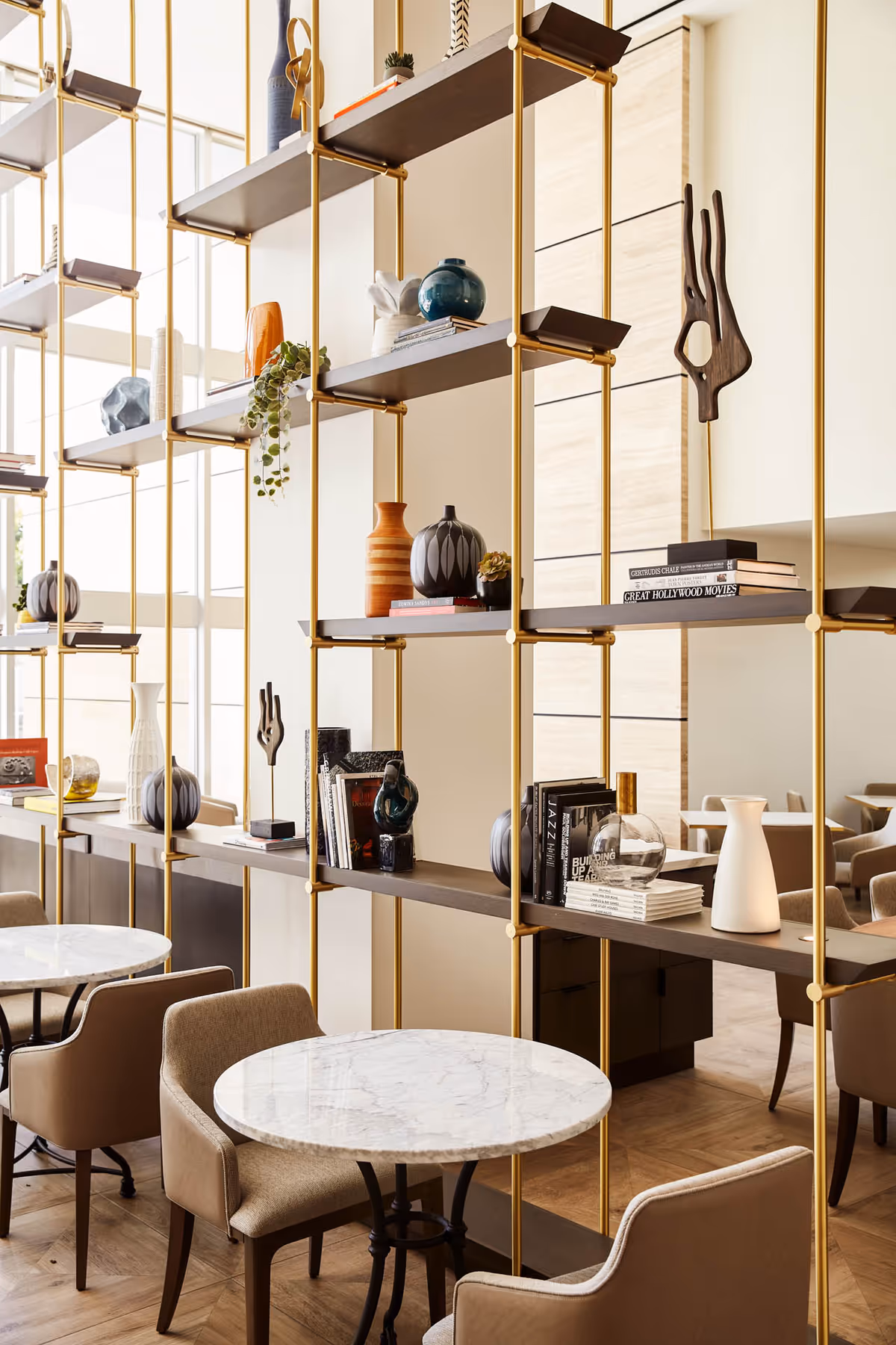 A modern interior space featuring round marble tables with beige upholstered chairs. A tall open shelving unit with gold metal supports and dark wooden shelves displays various decorative items including vases, books, and sculptures. The room has large windows allowing natural light to fill the space.