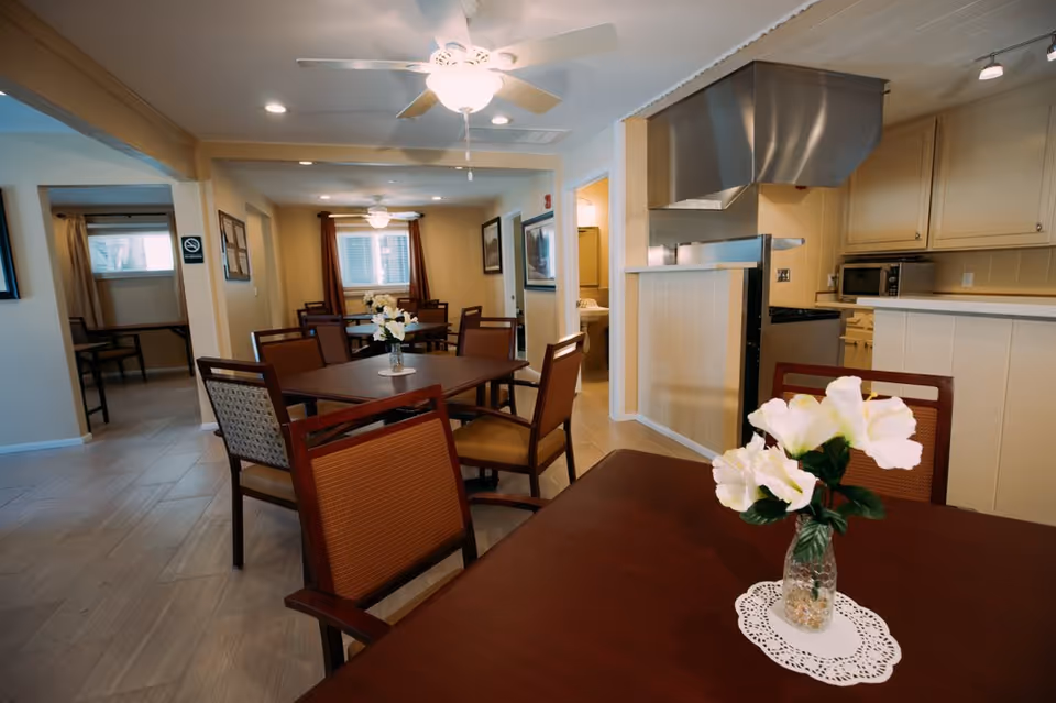 Dining room with multiple tables and chairs, a vase of flowers on the foreground table, and a kitchen pass-through in the background.