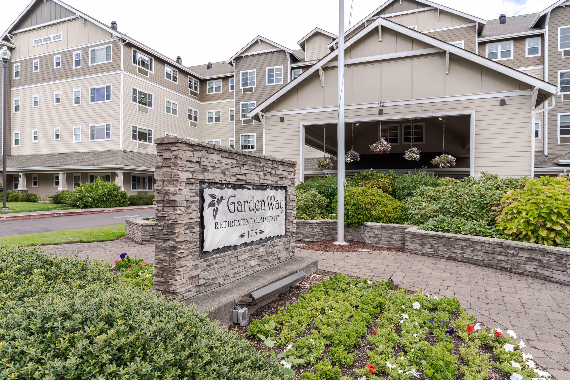 Exterior view of Garden Way Retirement Community building with a stone sign in the foreground displaying the community name and address. The building is multi-story with beige siding and numerous windows. There are landscaped bushes and flowers around the sign and along the walkway leading to the entrance.