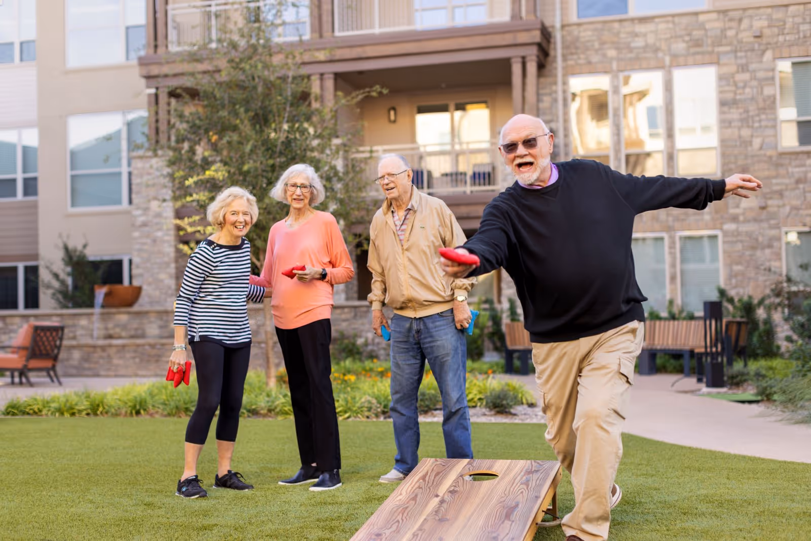 Four elderly people playing cornhole outside on a grassy area in front of a multi-story residential building. One man is in the act of throwing a bean bag, while the other three watch and smile. The setting appears to be a courtyard or garden area of a senior living community.