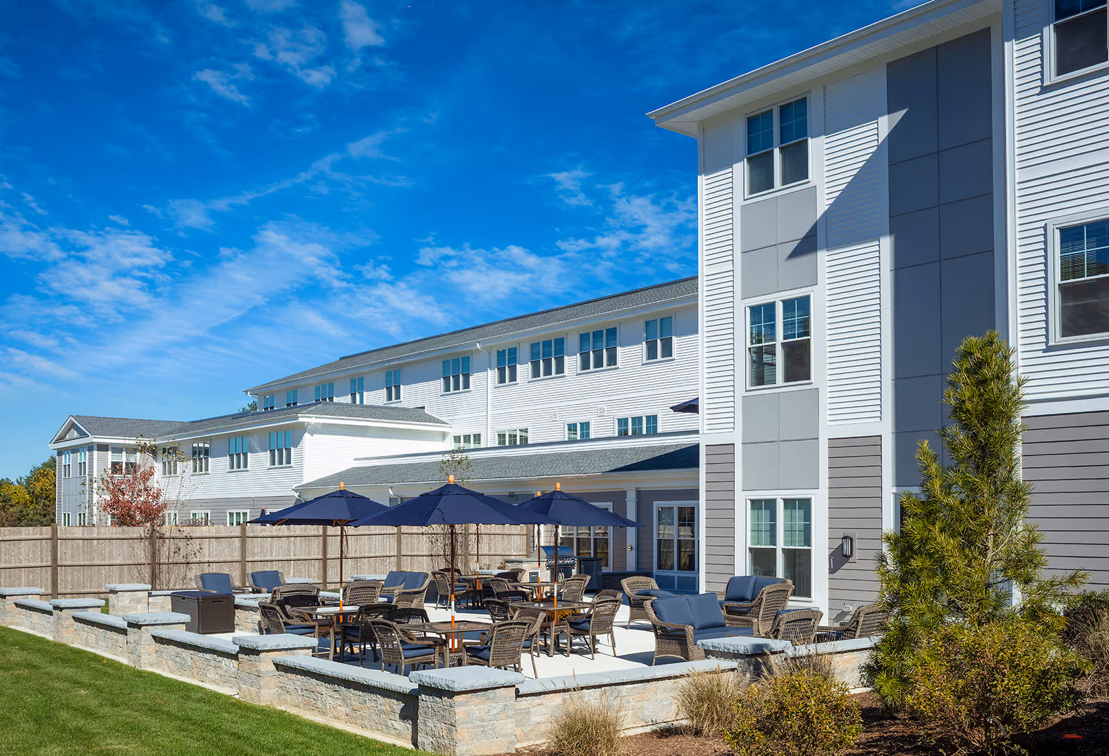 Outdoor patio area at Wingate Residences at Haverhill featuring multiple tables with blue umbrellas and wicker chairs, surrounded by a low stone wall, adjacent to a multi-story white and gray building under a bright blue sky.