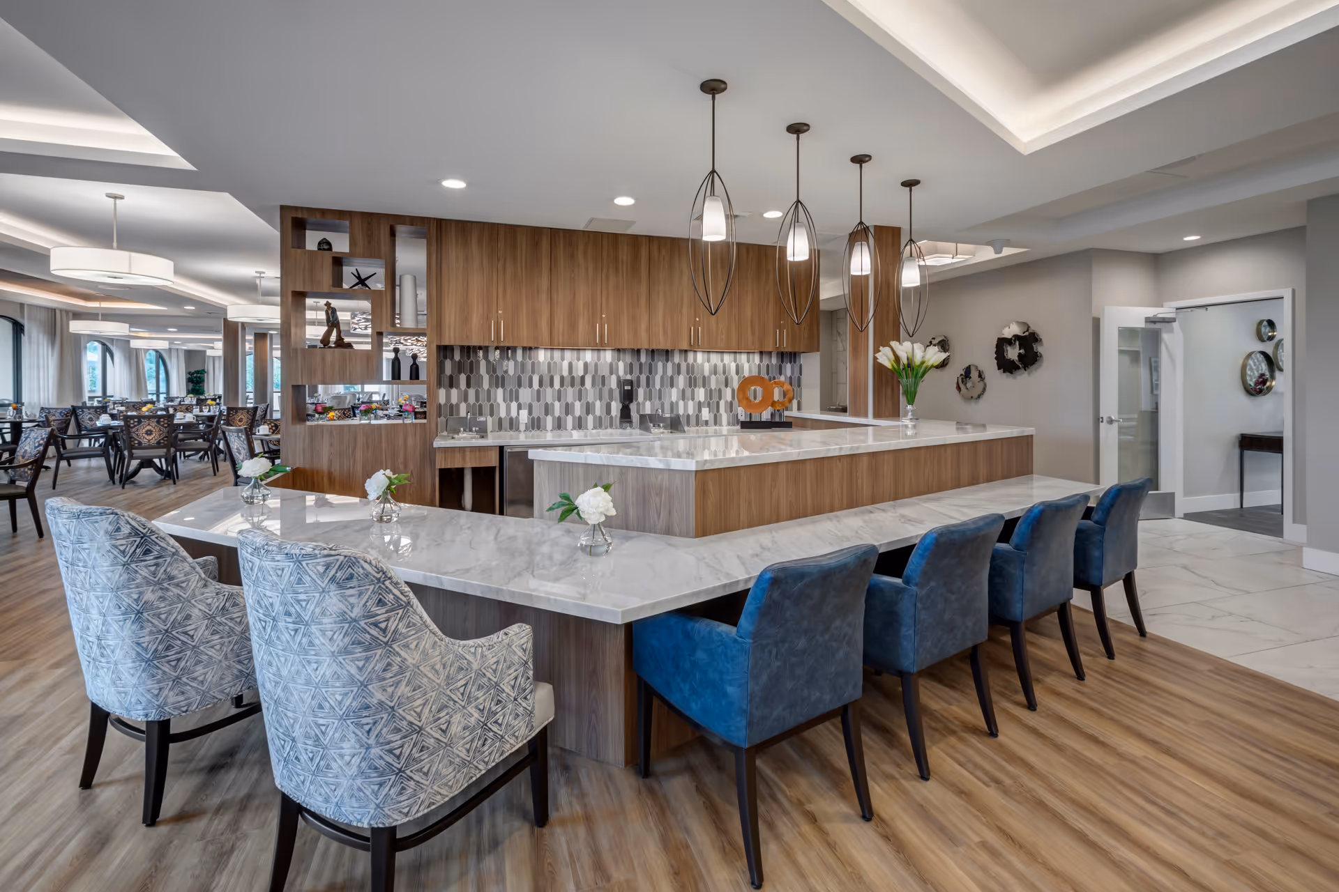 Modern dining area with a large marble countertop island surrounded by blue and patterned chairs. The background features wooden cabinetry with a geometric tile backsplash, pendant lights hanging from the ceiling, and a spacious dining room with multiple tables and chairs. The room has wood flooring and a bright, airy atmosphere.