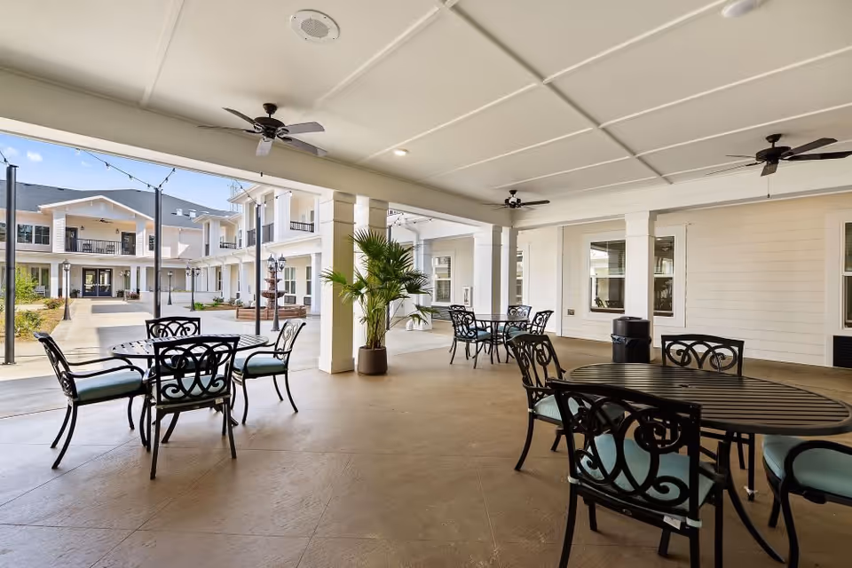 Covered outdoor patio area with several black metal tables and chairs with light blue cushions. Ceiling fans are mounted on the white ceiling, and there are potted plants near the columns. The patio overlooks a courtyard with a walkway, string lights, and a two-story building with balconies and windows.