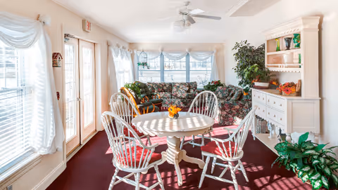 A bright and cozy living room area with large windows covered by white curtains allowing natural light to fill the space. The room features a round white table with four white chairs that have red cushions. In the background, there are two floral-patterned sofas, a white wooden cabinet with shelves holding decorative items, and several green plants adding a touch of nature to the room. The floor is covered with a dark red carpet, and a ceiling fan is mounted above.