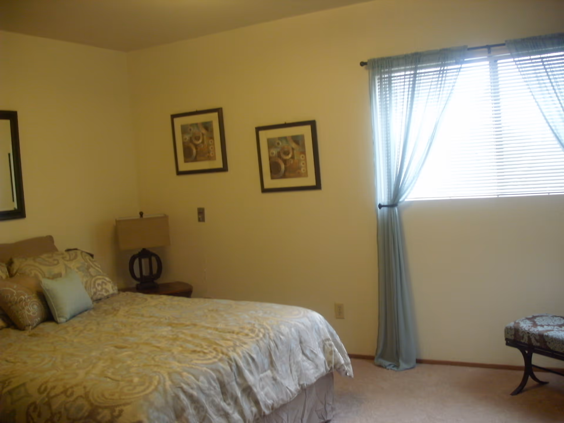 A bedroom with a bed covered in beige and light blue patterned bedding, a bedside table with a lamp, two framed pictures on the wall, a window with light blue curtains and blinds, and a cushioned bench near the window.