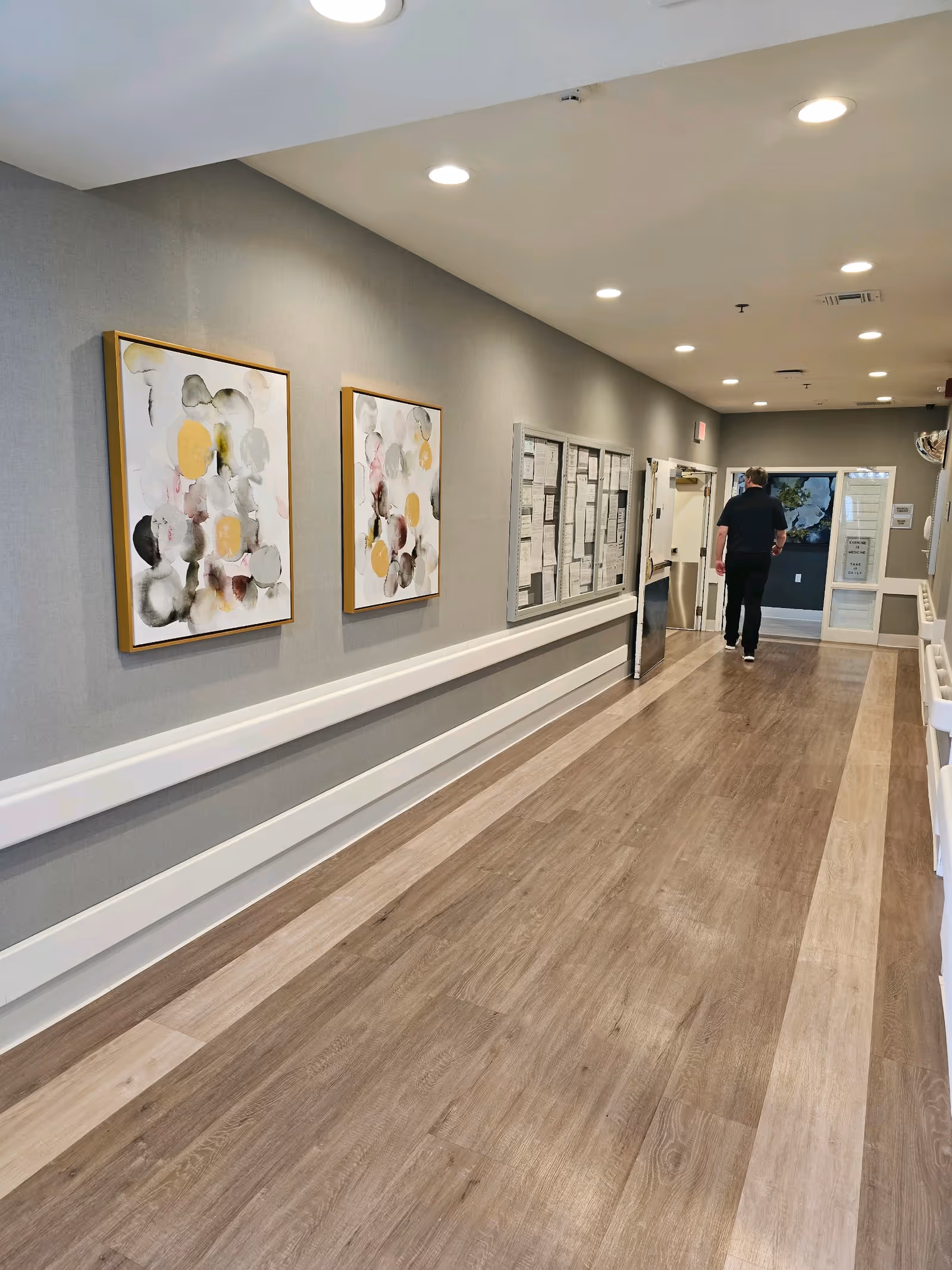 A long, well-lit hallway with wood-patterned flooring and gray walls. Two abstract paintings with circular patterns in muted colors are hung on the left wall above a white handrail. A bulletin board with notices is also visible on the left wall. A person dressed in black is walking away toward a door at the end of the hallway.