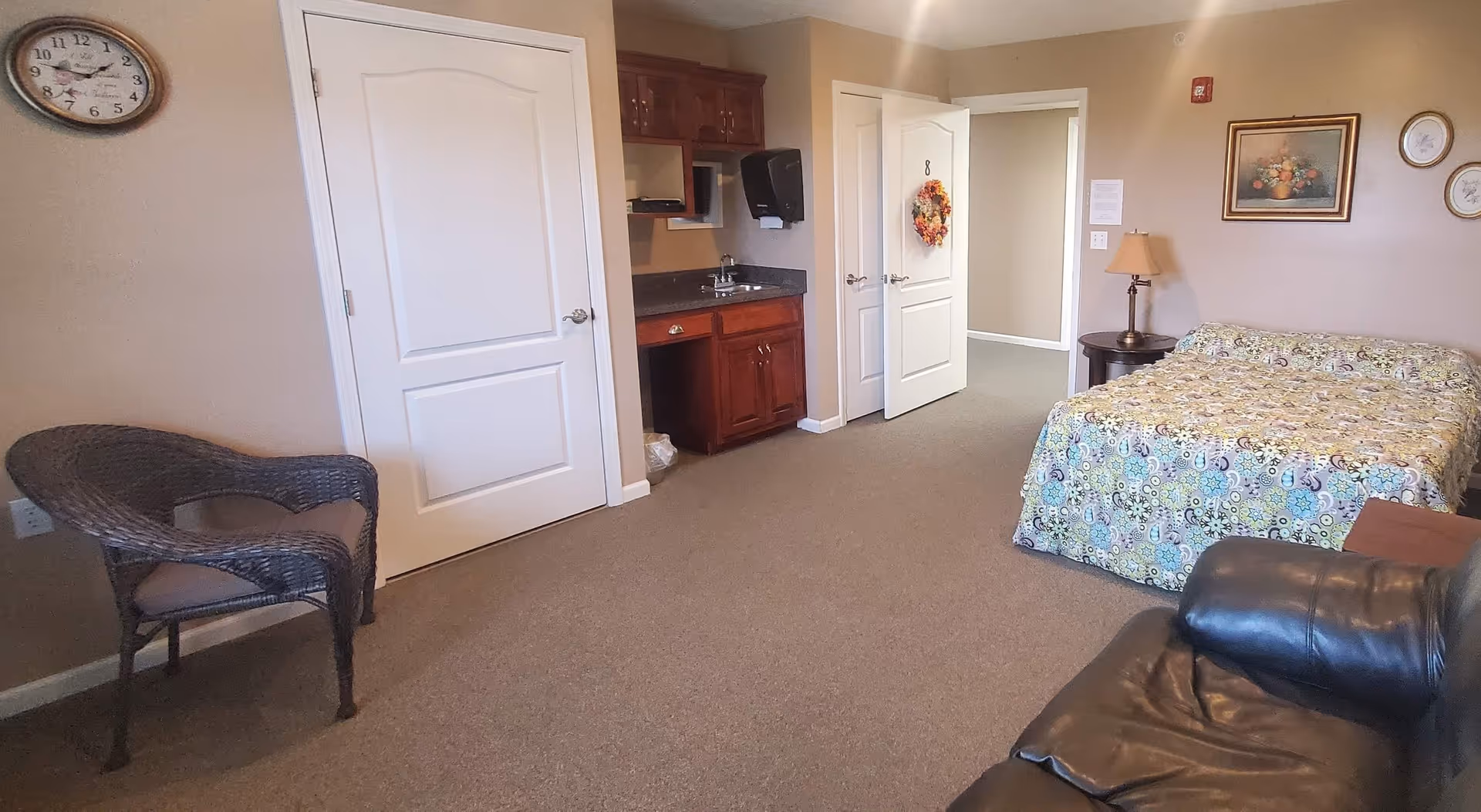 A residential care room with a bed covered in a colorful patterned bedspread, a black leather chair, a wicker chair with a cushion, a small wooden side table with a lamp, and a wall clock. There is a small kitchenette area with a sink and cabinets, and two white doors, one of which has a decorative wreath and the number 8 on it. The walls are beige and decorated with framed pictures.