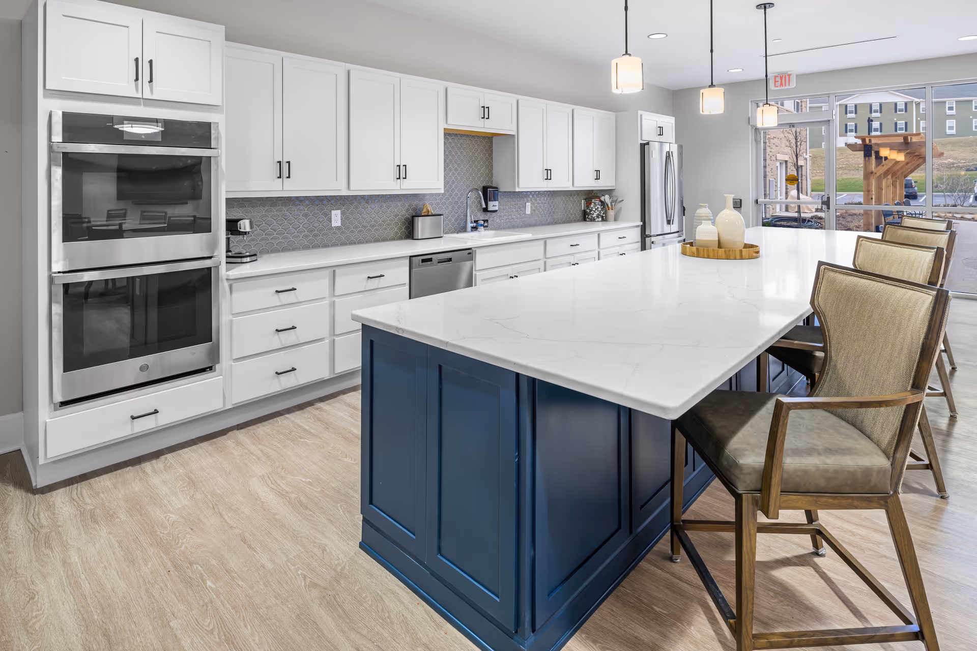Modern kitchen area with white cabinetry, stainless steel double oven, dishwasher, and refrigerator. A large island with a white marble countertop and blue base is surrounded by several cushioned chairs. Pendant lights hang above the island, and large windows in the background show an outdoor view with buildings and a wooden pergola.