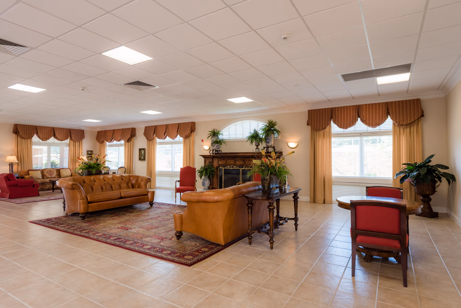 Spacious common room with leather sofas, armchairs, a fireplace, tables, and large windows with drapes.