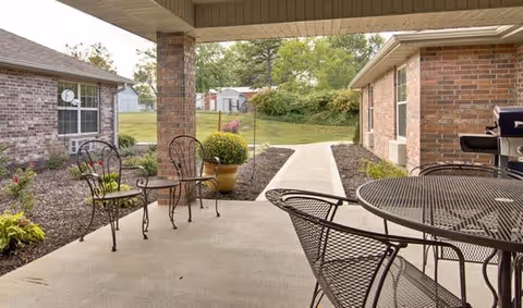 Covered outdoor patio area with metal chairs and tables, surrounded by brick buildings and landscaped garden beds with plants and flowers. A concrete walkway leads away from the patio into a grassy area with trees and a small shed in the background.
