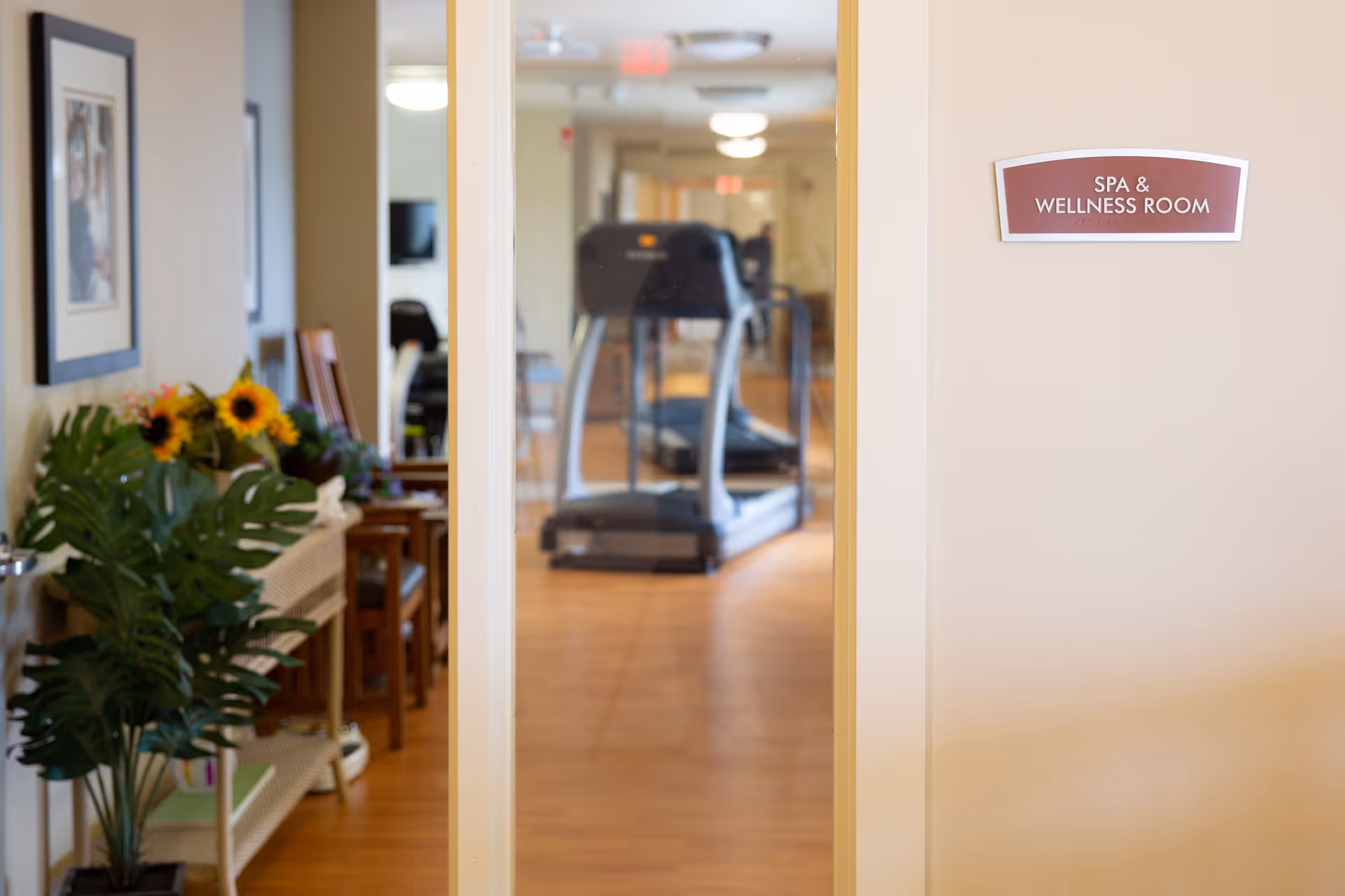 View through a doorway into a spa and wellness room with a treadmill visible inside. To the left of the doorway, there is a small table with a vase of sunflowers and a large green plant, along with some chairs and framed pictures on the wall.