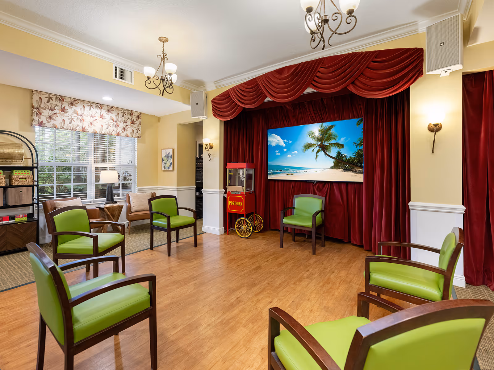 A cozy common area with six green cushioned chairs arranged in a circle on a wooden floor. The room features a large window with floral curtains, a popcorn machine, and a large screen displaying a beach scene with palm trees and blue sky. The walls are painted yellow with white wainscoting, and there are decorative light fixtures and red stage curtains framing the screen.