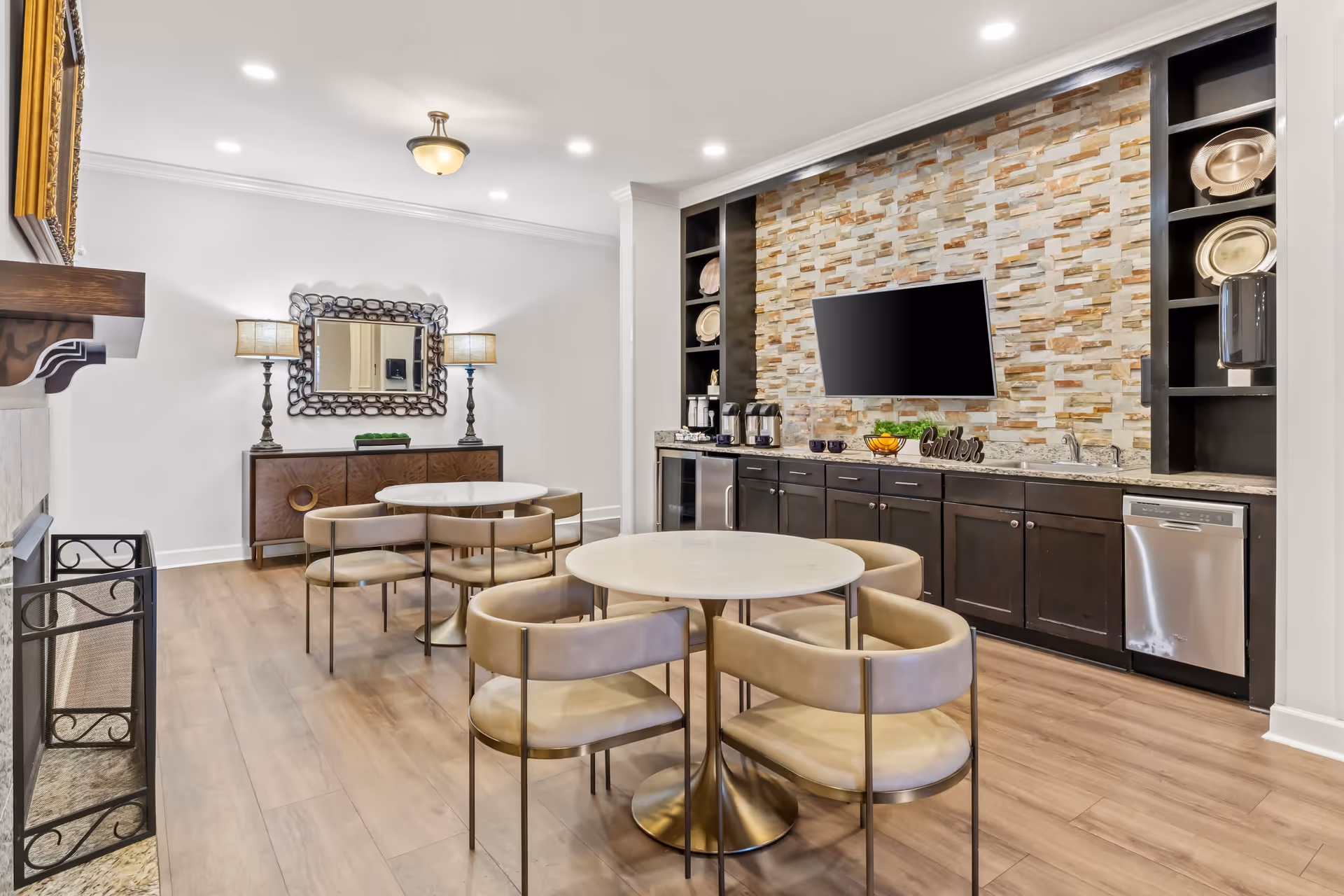 A modern common area with two round tables surrounded by beige cushioned chairs on a wooden floor. The back wall features dark cabinetry with a stone tile backsplash, a mounted flat-screen TV, and decorative plates on shelves. Two table lamps flank a decorative mirror above a wooden sideboard. The room is well-lit with recessed ceiling lights and a central ceiling fixture.