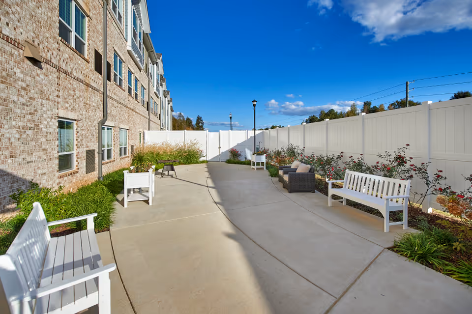 Outdoor patio area at a senior living facility with white benches, a wicker loveseat with cushions, planters, and a concrete walkway. The area is enclosed by a white fence and the building's brick exterior is visible on the left side under a blue sky with some clouds.