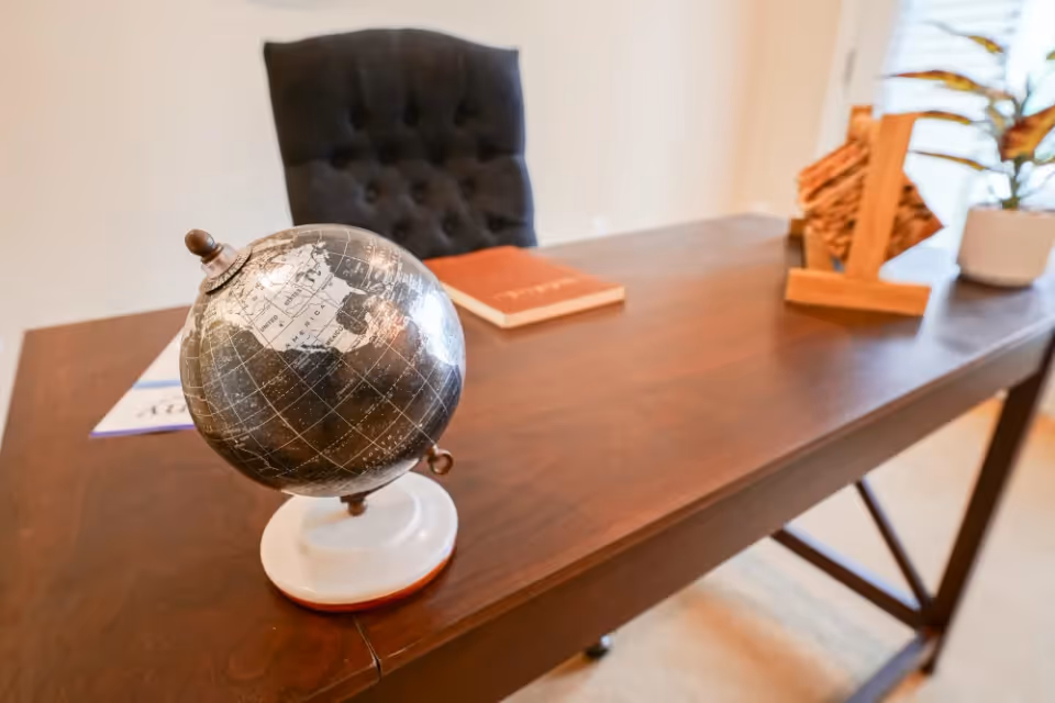 A close-up view of a wooden desk with a small decorative globe, a book, a wooden card holder, and a potted plant. A dark tufted chair is positioned behind the desk.