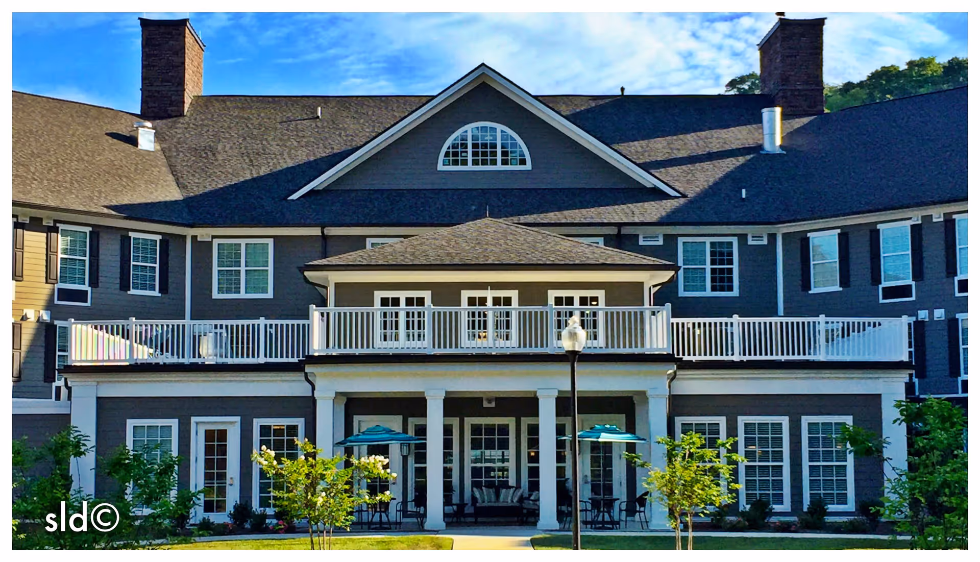 Exterior view of a senior living facility building with gray siding, white trim, multiple windows, and two chimneys. The building features a covered porch with white columns, outdoor seating with umbrellas, and a balcony above. There is a lamp post and small trees in the landscaped front yard under a blue sky with some clouds.