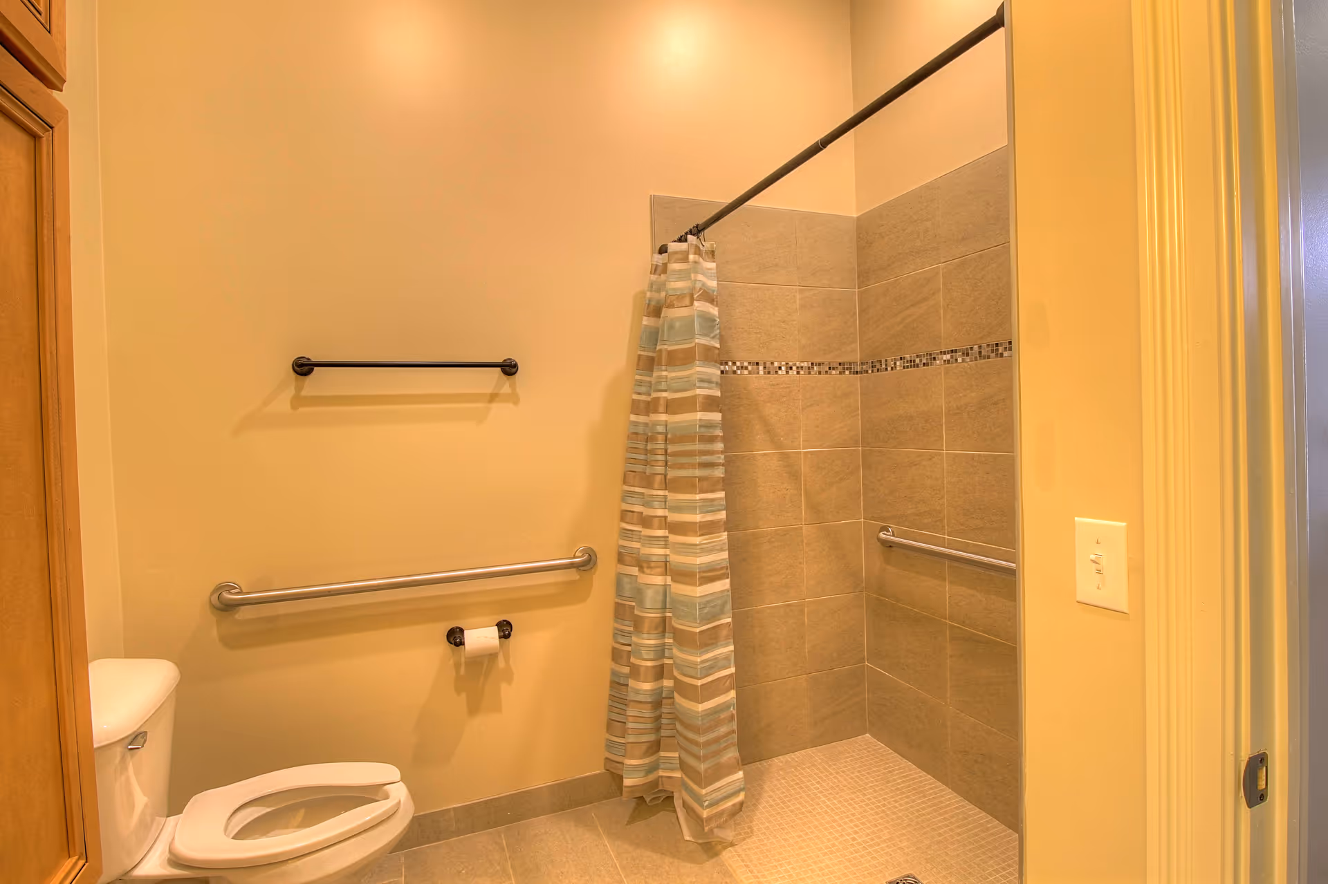 A bathroom with a toilet, a wall-mounted toilet paper holder, two grab bars, a towel rack, and a walk-in shower with a striped shower curtain and tiled walls.