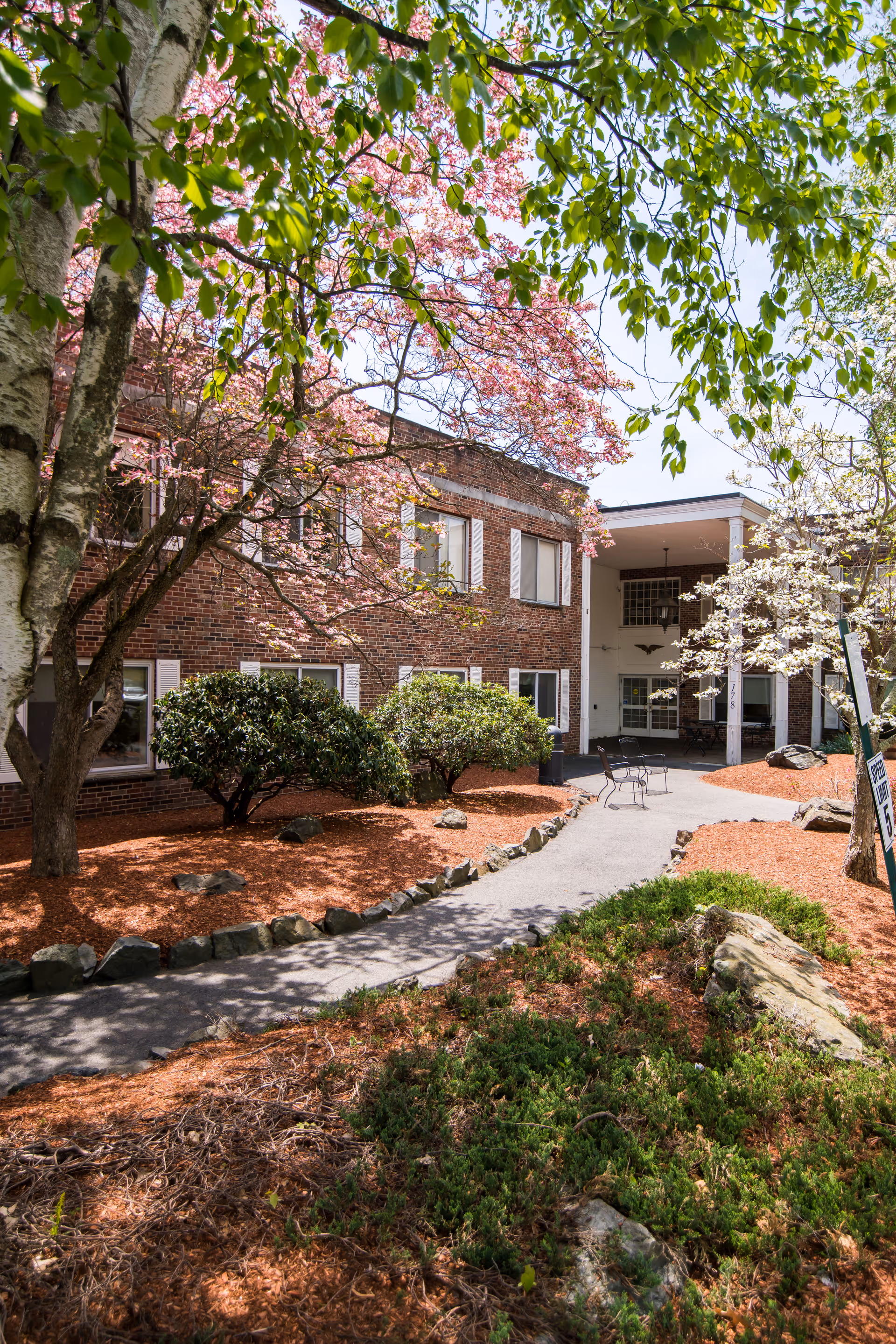Outdoor view of a brick building with white window shutters surrounded by landscaped garden beds with mulch, rocks, bushes, and blooming trees with pink and white flowers. A paved walkway leads to a covered entrance with outdoor seating.