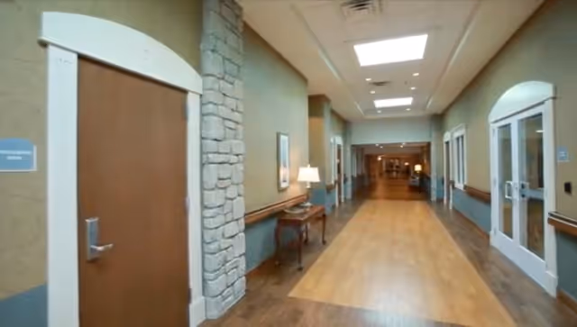 A wide hallway in a senior living community with wooden floors, beige walls, and stone accents. There are doors on both sides, a small table with a lamp and framed artwork on the left wall, and ceiling lights illuminating the corridor.