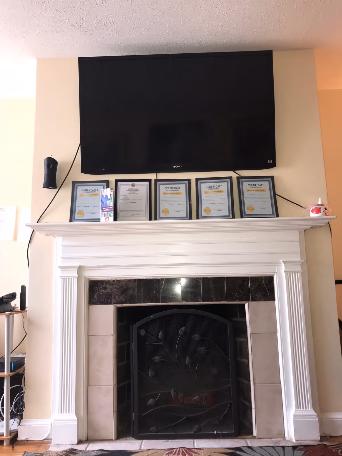 A flat-screen TV mounted above a white fireplace mantel lined with framed certificates.