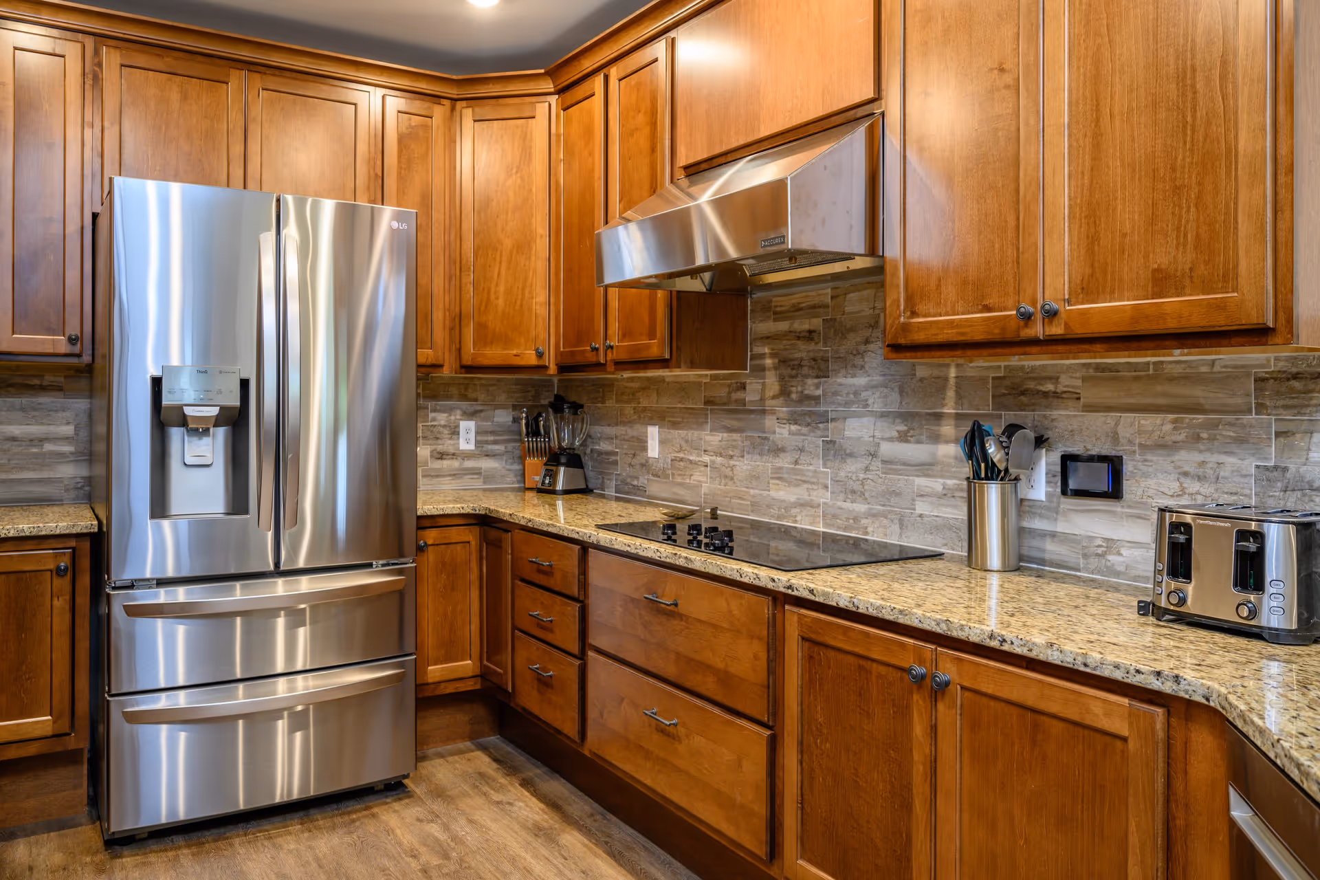 Modern kitchen with wooden cabinets, a stainless steel refrigerator, a built-in stovetop with a stainless steel range hood, a blender, a utensil holder, and a toaster on granite countertops with a tiled backsplash.
