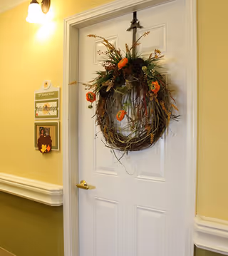 A white door decorated with a fall-themed wreath featuring orange flowers and greenery. The door is in a hallway with yellow walls and white trim. There is a light fixture above the door and a small sign with a name and a picture of two people on the wall next to the door.