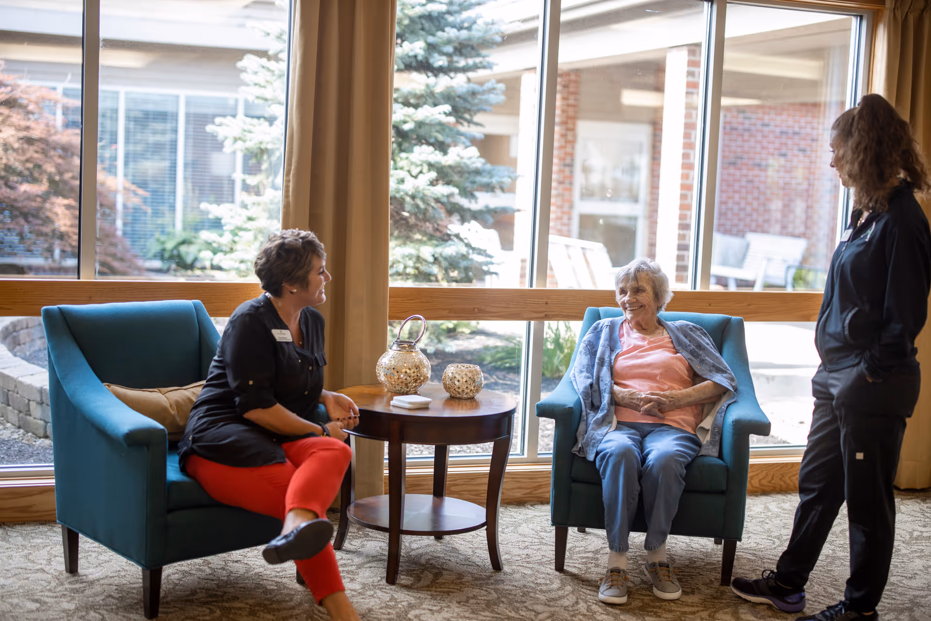 An elderly woman sitting in a blue armchair smiling and talking with two caregivers in a bright room with large windows showing an outdoor garden area. There is a small round wooden table between the chairs with decorative items on it.